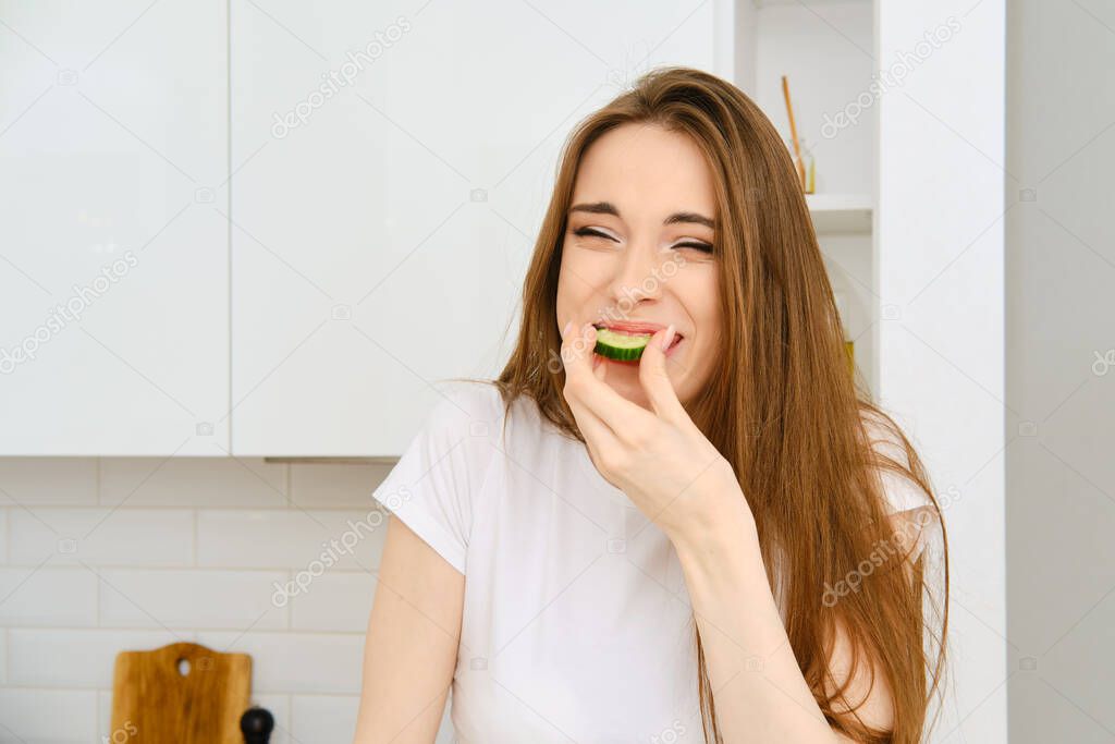 Funny young joyful woman in white t-shirt bites slice of cucumber, squints and smiling. Happy female having fun while cooking in modern white kitchen at home
