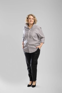 Gri gömlekli ve pantolonlu yaşlı bir kadının gri stüdyo arkaplanı üzerindeki tüm vücut portresi.