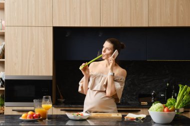 Hamile kadın, mutfakta yemek hazırlarken kereviz sapını kemiriyor ve telefonla konuşuyor.