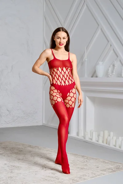 Beautiful young woman in full body red fishnet bodysuit posing in studio with one hand on waist