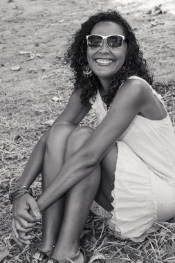 Siyah model, güneş gözlüklü, siyah beyaz, parkta çimlerin üzerinde oturan, gülümseyen ve kameraya bakan. Neşe kavramı, olgunluk ve siyah farkındalık. Salvador, Bahia, Brezilya.