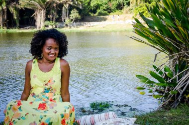 Baskın yeşil elbiseli kadın ağaçların, bitkilerin, çiçeklerin ve arka plandaki nehrin arasında fotoğraf için poz veriyor. Neşe kavramı, olgunluk ve siyah farkındalık.