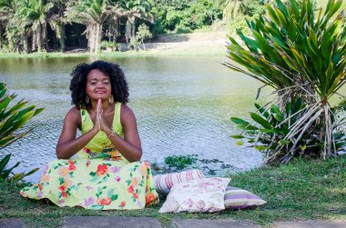 Baskın yeşil elbiseli kadın ağaçların, bitkilerin, çiçeklerin ve arka plandaki nehrin arasında fotoğraf için poz veriyor. Neşe kavramı, olgunluk ve siyah farkındalık.