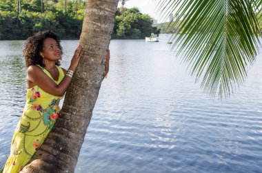 Siyah kadın fotoğraf için poz veriyor. Gülümsüyor ve kameraya bakıyor. Arka plandaki ağaçlar, nehir ve bitkiler. Salvador, Bahia, Brezilya.