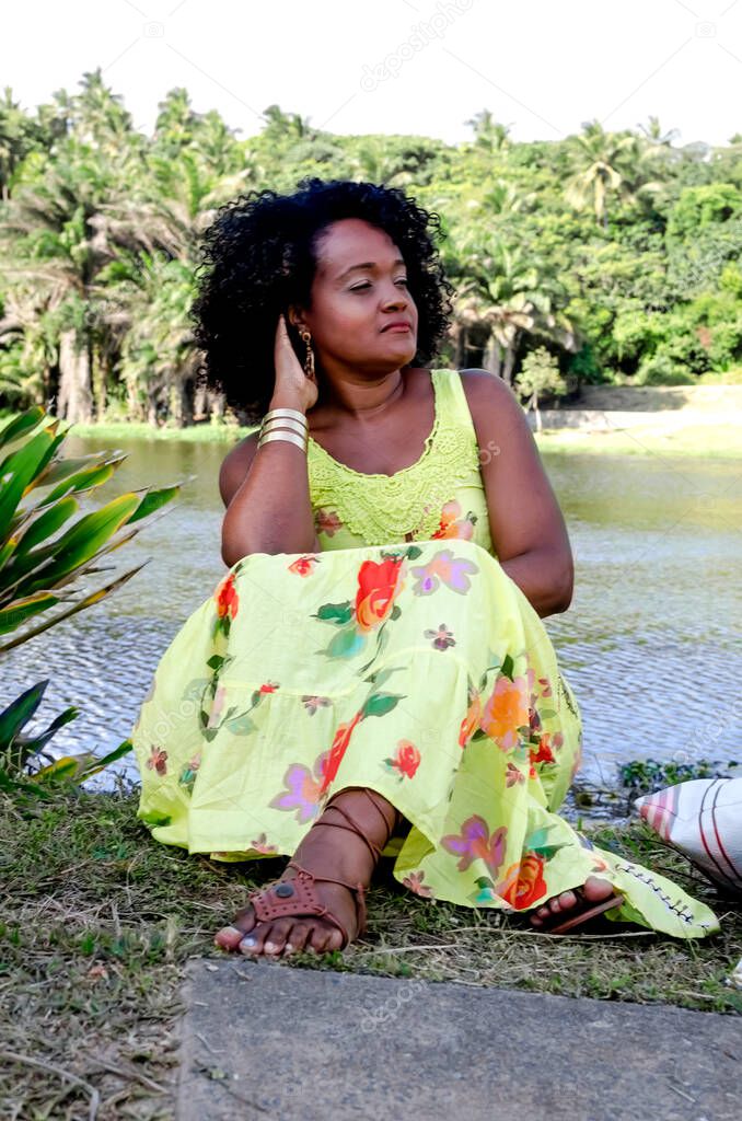 Woman with predominant green colored dress posing for photo among trees plants and flowers and river in background. Concept of joy, maturity and black awareness.