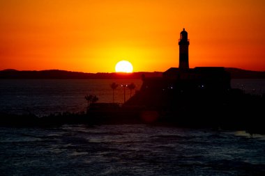 Salvador, Bahia, Brezilya - 19 Eylül 2021: Brezilya 'nın Bahia eyaletindeki Barra plajında güzel ve dramatik bir turuncu gün batımına karşı bir deniz fenerinin silueti.