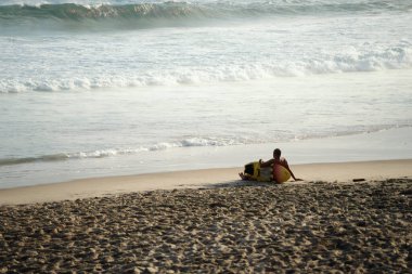 Salvador, Bahia, Brezilya - 26 Eylül 2021: Sahilde oturmuş denize bakan sörfçü. Farol da Barra plajı Salvador, Bahia eyaleti, Brezilya.