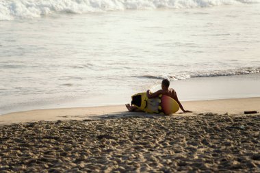 Salvador, Bahia, Brezilya - 26 Eylül 2021: Sahilde oturmuş denize bakan sörfçü. Farol da Barra plajı Salvador, Bahia eyaleti, Brezilya.