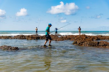 Salvador, Bahia, Brezilya - 10 Ekim 2021 Rio Vermelho sahilindeki kayalıklarda balık tutan erkekler sırıkla, Bahia, Brezilya 'nın başkenti Salvador' da balık tutuyorlar.