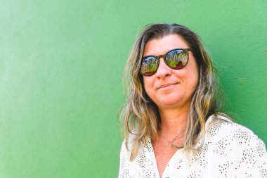 Güneş gözlüğü takan bir kadının portresi yeşil bir evin duvarına yaslanmış. Maragogipinho, Bahia, Brezilya.