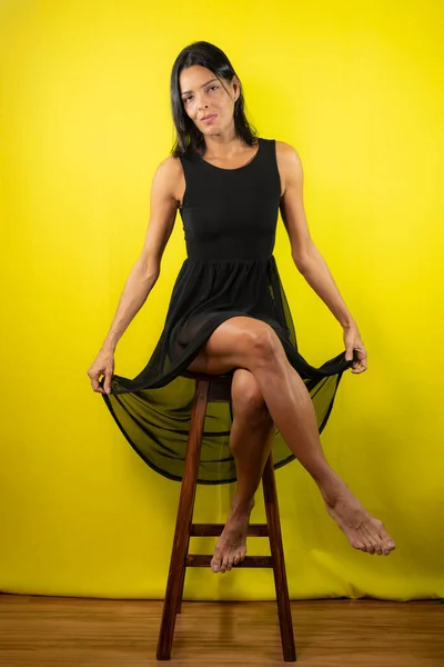 Portrét Modelu Sedícího Dřevěné Lavičce Držícího Černé Šaty Žlutém Pozadí — Stock fotografie