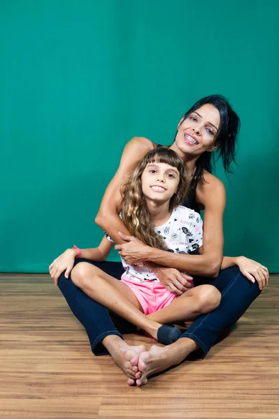 Portrait of mother and daughter sitting on wooden floor against dark green background. Salvador, Bahia, Brazil.