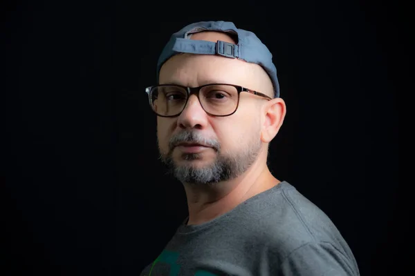 Portrait of a bald bearded man wearing cap and glasses looking at the camera against a black background. Salvador, Bahia, Brazil.