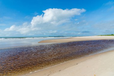 Deniz ve nehir ıssız sahilde, bulutlu mavi gökyüzüne karşı buluşuyor. Praia do Guaibim, Brezilya Bahia Denizi kıyısında.