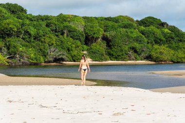 Genç ve güzel bir kadın kumsalda ağaçlara ve mangrove ağaçlarına doğru yürüyor. Praia do Guaibim, Brezilya Bahia Denizi kıyısında.