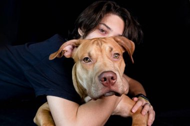 Siyah arka planda karamel renkli pitbull köpeği olan genç bir adamın portresi. Salvador, Bahia, Brezilya.