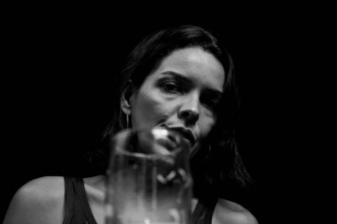 Studio Portrait in black and white of young woman in black t-shirt drinking beer from glass cup. against black studio background. Salvador, Brazil.