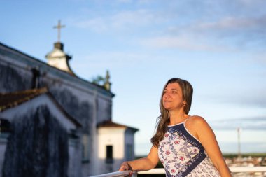 A woman on the porch of her house in light clothes looking at the street against the sky and church in the background. 