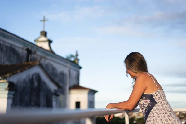 A woman on the porch of her house in light clothes looking at the street against the sky and church in the background.