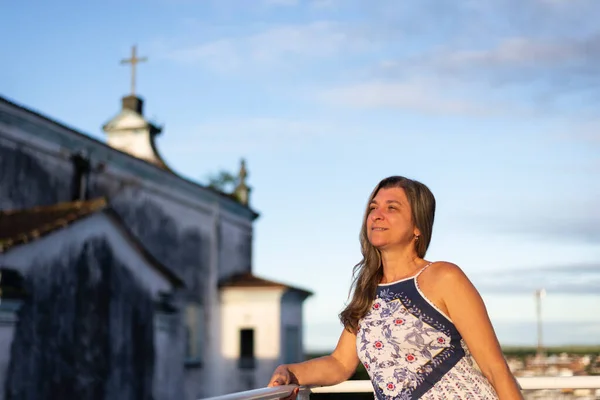 A woman on the porch of her house in light clothes looking at the street against the sky and church in the background.