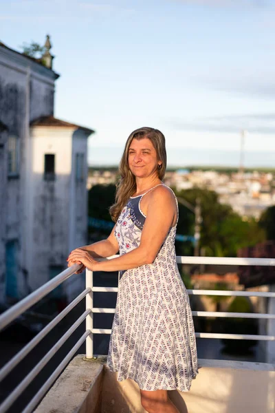 A woman on the porch of her house looking at the camera against the sky and church in the background.