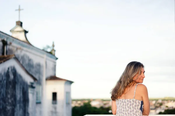 A woman on the porch with her back to the camera against the sky and church in the background.