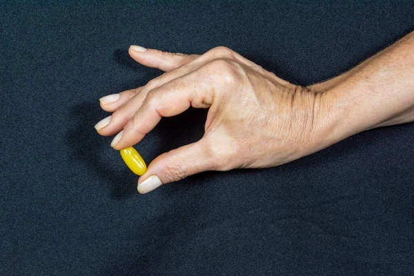 A yellow pill held by the fingers of one hand against a black background. Medical material.
