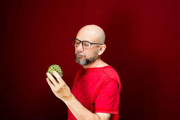 Young handsome man with bald beard wearing glasses and red shirt standing holding pinecone fruit against red background. Positive and healthy person.