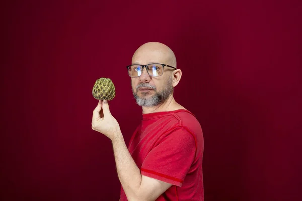Young handsome man with bald beard wearing glasses and red shirt standing holding pinecone fruit against red background. Positive and healthy person.