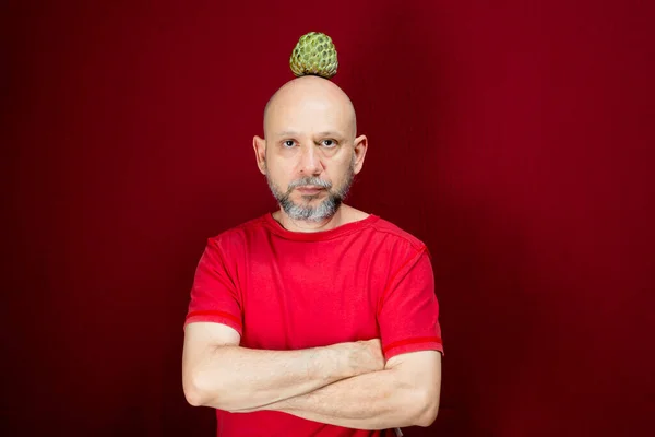 Young handsome man with beard, bald head and red shirt standing with pinecone fruit balanced on head against red background. Positive and healthy person.