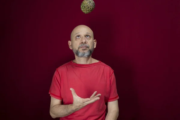Young handsome man with beard, bald head and red shirt standing tossing pinecone fruit up against red background. Positive and healthy person.