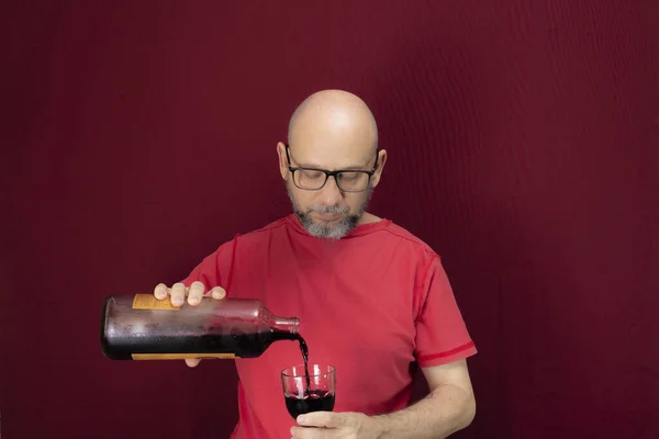 Young handsome man with beard, bald, red shirt wearing glasses holding in his hands a bottle of grape juice and a glass glass against red background. Positive and healthy person.