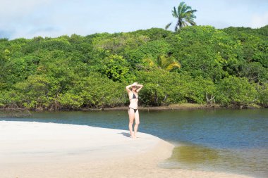 A woman, in a black bikini, is on the sand next to the red water river against the green forest in the background. Praia do Guaibim, coast of the sea of Bahia, Brazil