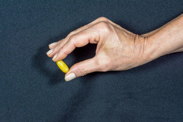 A yellow pill held by the fingers of one hand against a black background. Medical material.