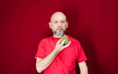 Young handsome man with beard, bald head and red shirt standing holding pinecone fruit in hand against red background. Positive and healthy person.