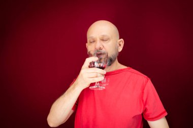Handsome man with beard, bald man in red shirt enjoying the taste of grape juice in glass cup against red background. Healthy and cheerful person.