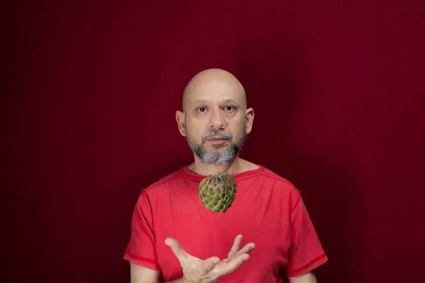 Young handsome man with beard, bald head and red shirt standing tossing pinecone fruit up against red background. Positive and healthy person.