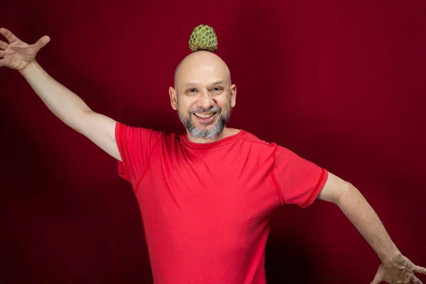 Young handsome man with beard, bald head and red shirt standing with pinecone fruit balanced on head against red background. Positive and healthy person.