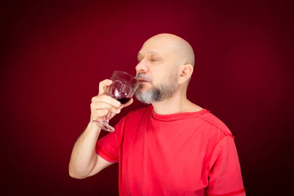 Handsome man with beard, bald man in red shirt enjoying the taste of grape juice in glass cup against red background. Healthy and cheerful person.