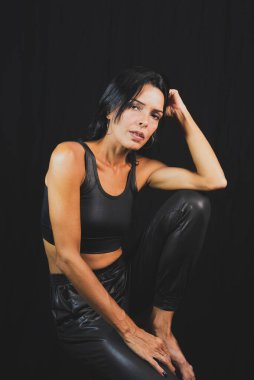 Studio portrait of young woman in black t-shirt and pants sitting on wooden bench against black studio background. Salvador, Brazil.