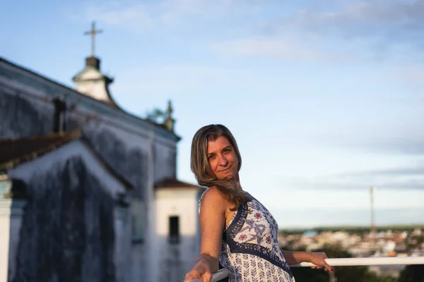 A woman on the porch of her house looking at the camera against the sky and church in the background. City of Valenca, Bahia, Brazil.