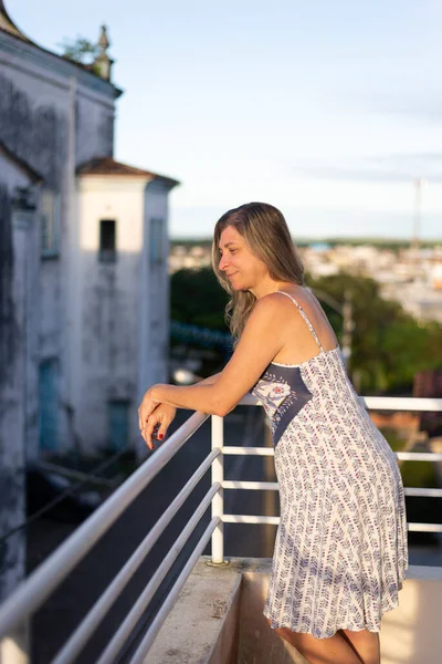 A woman on the porch of her house in light clothes looking at the street against the sky and church in the background. City of Valenca, Bahia, Brazil.