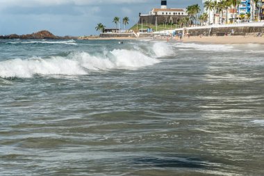 Low view of a beach against blue sky and buildings in the background. Barra beach lighthouse, Salvador, Brazil.