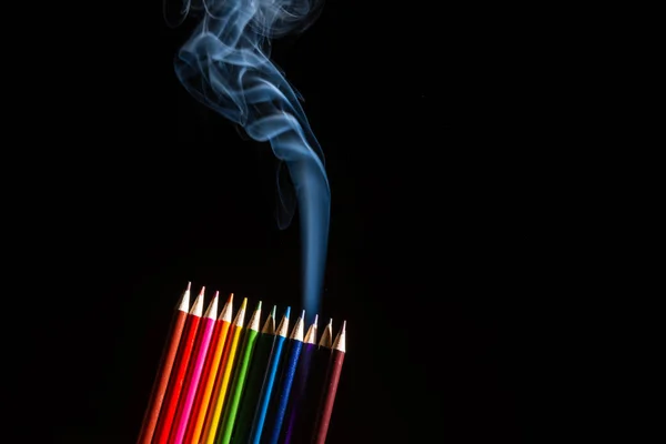 Colored pencils grouped on black background with white smoke coming from below. Close up. Sharp pencil scraps.