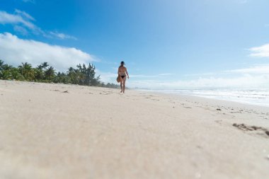 An adult woman in a bikini walking under strong sunlight on Guaibim beach in the city of Valenca, Brazilian state of Bahia. Travel and fun.
