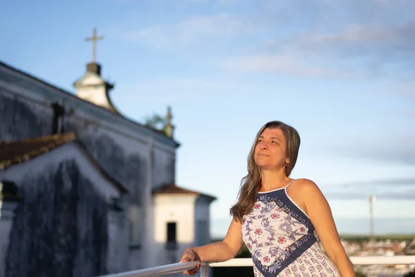 A woman on the porch of her house in light clothes looking at the street against the sky and church in the background. City of Valenca, Bahia, Brazil.