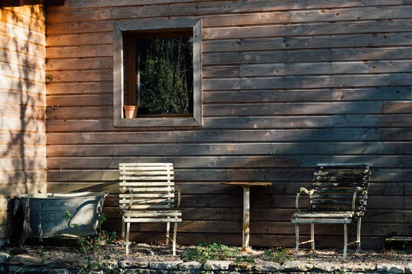 Ahşap bir evin önündeki iki eski demir koltuğun manzarası. Bir barakanın önündeki sakin sahne.