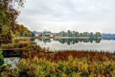 Ağaçların, çan kulesinin, küçük bir ahşap iskelenin ve bataklık sazlıkların yansıtıldığı Pusiano Gölü 'nün sonbahar manzarası..