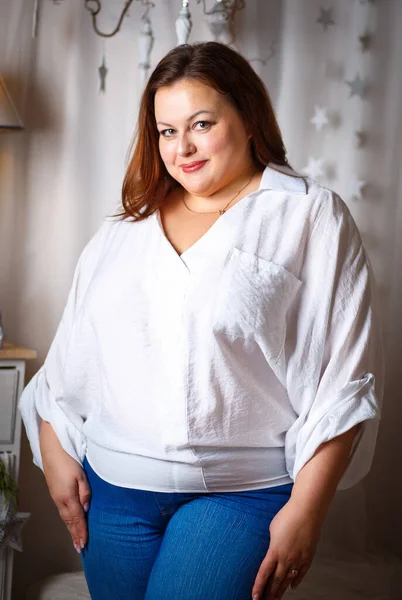 Beyaz bluz ve kot pantolon giyen güzel, büyük beden bir kız hafif bir Noel arka planında duruyor.