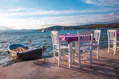 Boş mor kaplı beyaz masa, deniz kenarında oturmaya hazır. Tekne masanın yanında bekliyor. Huzurlu, deniz manzaralı sessiz bir masa. Yeşil iklimi destekler.
